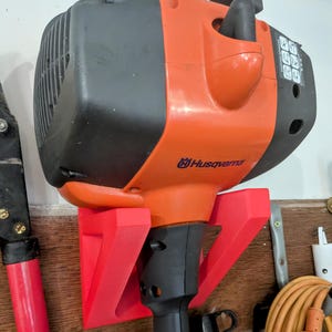 May include: An orange and black Husqvarna string trimmer mounted on a red bracket. The trimmer is attached to a wooden wall. The image shows the trimmer's engine and handle, with the Husqvarna logo visible.