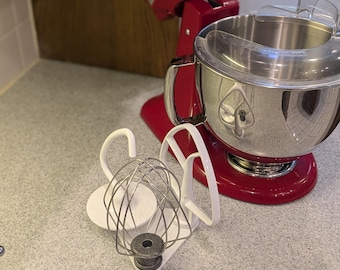 Op de afbeelding: Een rode keukenmixer met een roestvrijstalen kom en diverse opzetstukken, waaronder een garde en een platte klopper. De mixer staat op een gespikkelde aanrecht, een keukenapparaat.