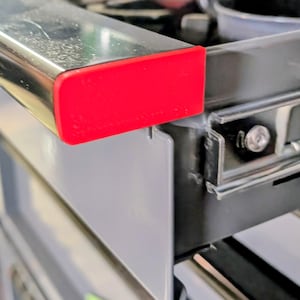 May include: A metallic drawer handle featuring a bright red rectangular end cap. The handle is attached to a grey drawer front with a visible metal sliding rail mechanism and a small silver screw securing the assembly to the drawer frame.
