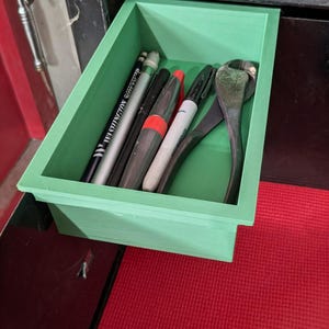 May include: A light green rectangular drawer filled with various tools and writing utensils. Items include pencils, markers, and a wrench. The drawer is set against a dark background with a red surface.