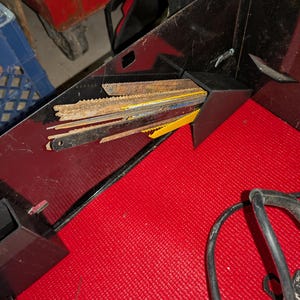 May include: A collection of various metal saw blades, some with yellow and blue accents, stored in a black container. The blades are of different sizes and show signs of use. The container sits on a red textured surface.