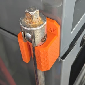 May include: An orange 3D-printed tool holder with a honeycomb pattern, holding a metal socket wrench. The holder is mounted on a gray surface, with the number "4F" printed on the side. The wrench has signs of rust.