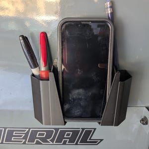 May include: A black and gray phone and pen holder with a smartphone, a black marker, a red marker, and a blue pencil. The holder is mounted on a gray surface.