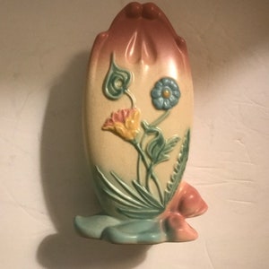 Puede incluir: Un jarrón de pared vintage con forma de tulipán. El jarrón es principalmente de color crema con un degradado rosa y melocotón en la parte superior. Presenta diseños florales en relieve en amarillo, azul y verde. La base es azul claro.