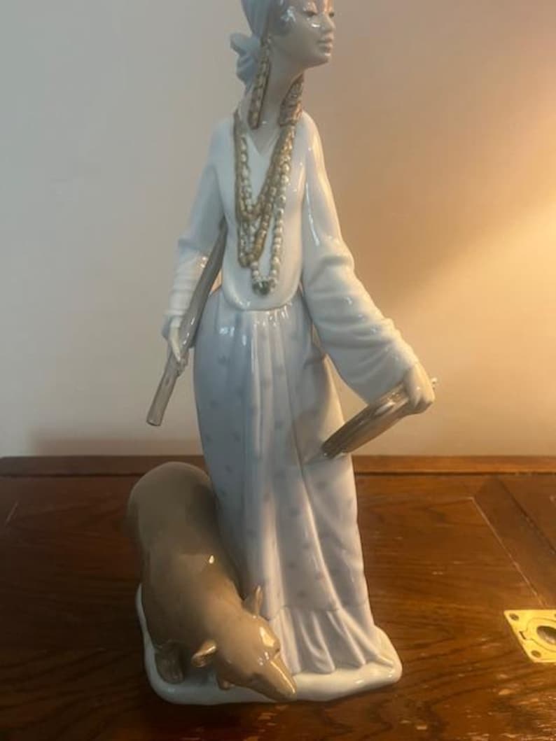 May include: A porcelain figurine of a woman in a blue and white dress with a brown bear at her feet. She is wearing a head scarf and multiple necklaces.