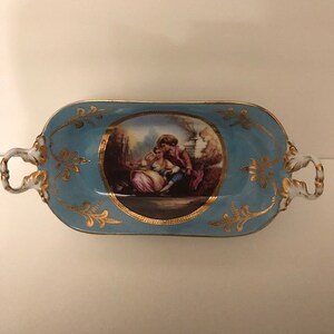 May include: A blue and gold porcelain serving dish with a painted scene of a couple in a garden. The dish has two handles and a gold rim.