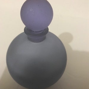 May include: A frosted glass bottle with a round, light purple stopper. The bottle is a light blue colour and has a smooth, rounded shape.