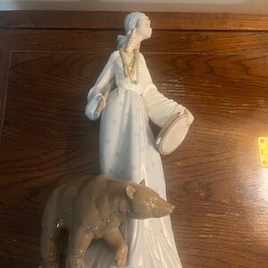 May include: A porcelain figurine of a woman in a white dress holding a tambourine and a brown bear. The woman is wearing a beaded necklace and a head scarf.