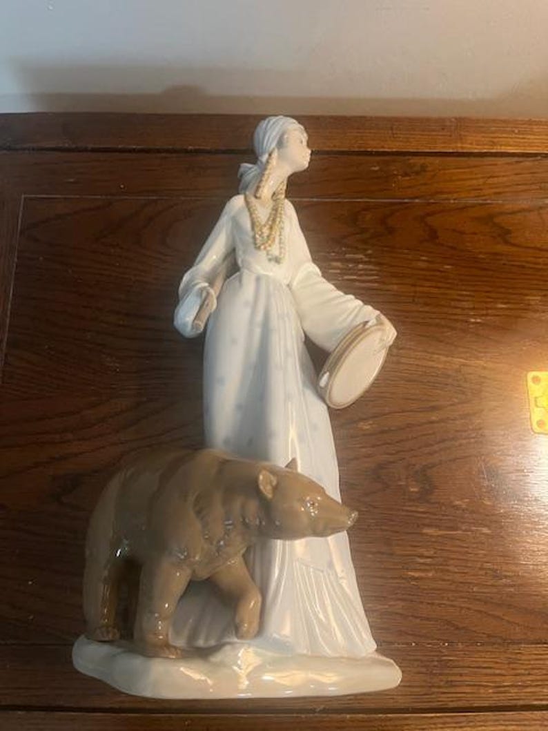 May include: A porcelain figurine of a woman in a white dress holding a tambourine and a brown bear. The woman is wearing a beaded necklace and a head scarf.
