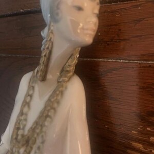 May include: A porcelain figurine of a woman wearing a white headscarf and a long white dress with gold accents. She has a delicate facial expression and is wearing large gold earrings.