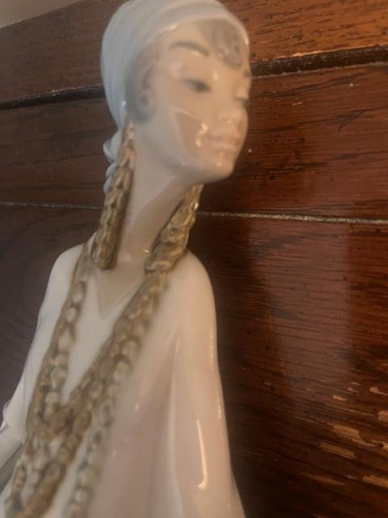 May include: A porcelain figurine of a woman wearing a white headscarf and a long white dress with gold accents. She has a delicate facial expression and is wearing large gold earrings.