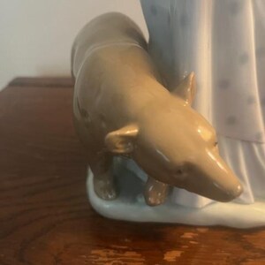 May include: A porcelain figurine of a woman in a white dress with blue polka dots, standing next to a brown dog. The figurine is on a white base.