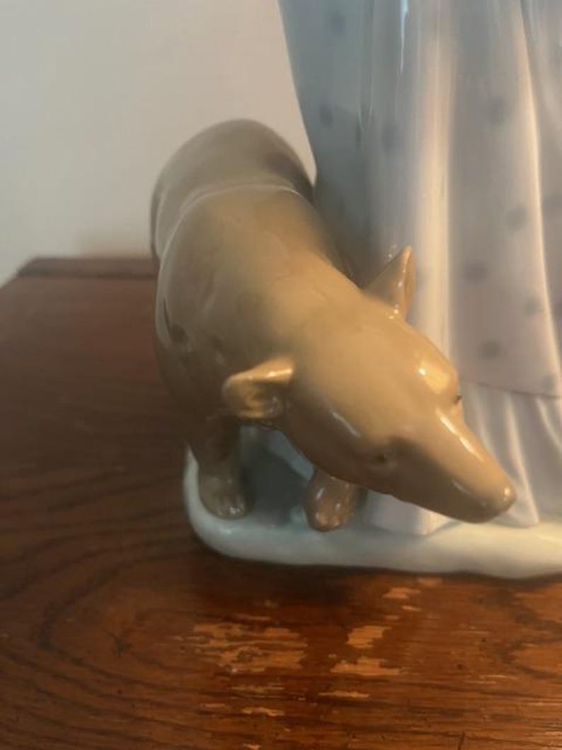May include: A porcelain figurine of a woman in a white dress with blue polka dots, standing next to a brown dog. The figurine is on a white base.