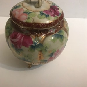 May include: A vintage porcelain jar with a lid and three feet. The jar is decorated with a floral pattern of pink and red roses, green leaves, and gold accents.