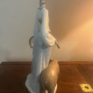 May include: A porcelain figurine of a woman in a blue and white dress holding a staff and a drum. She is standing next to a brown pig.