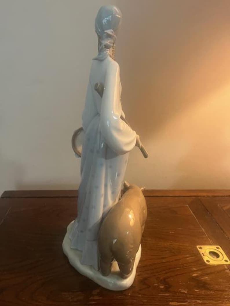 May include: A porcelain figurine of a woman in a blue and white dress holding a staff and a drum. She is standing next to a brown pig.