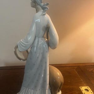 May include: A porcelain figurine of a woman in a blue and white dress holding a tambourine and walking with a brown animal.