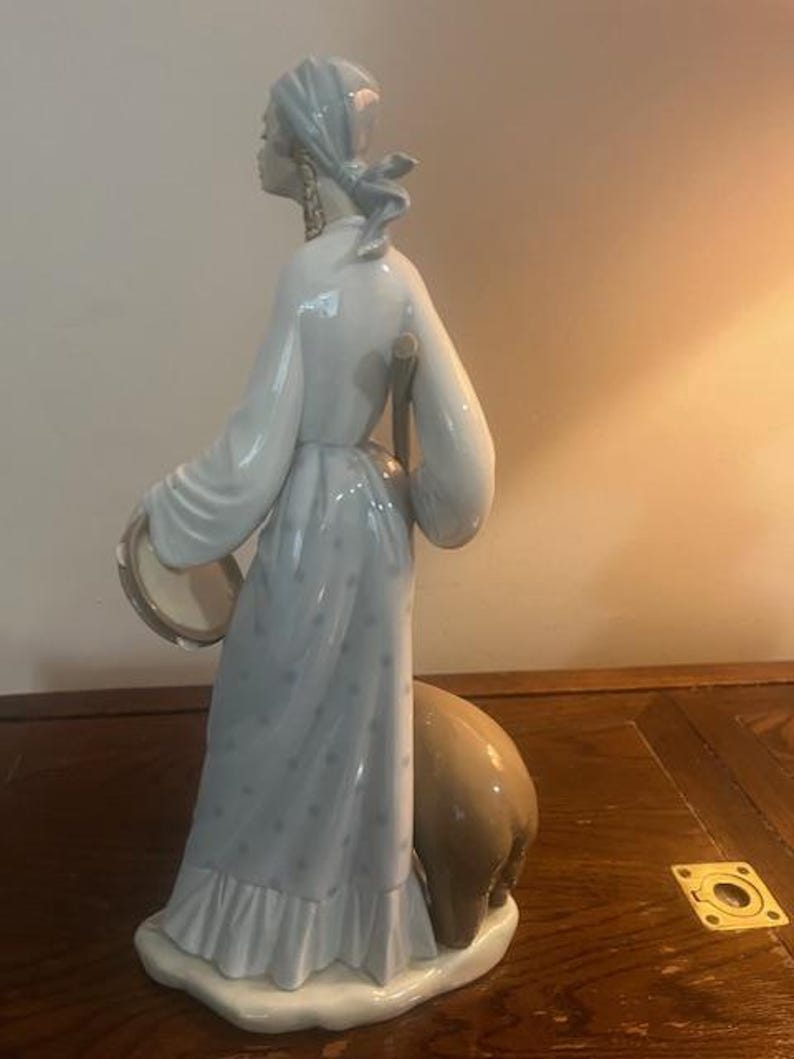 May include: A porcelain figurine of a woman in a blue and white dress holding a tambourine and walking with a brown animal.