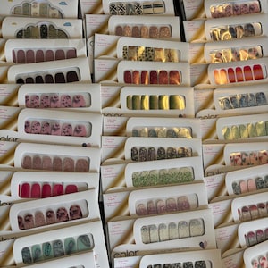 May include: A collection of Color Street nail polish strips in various designs and colors. The nail strips are packaged in white cardboard containers, showcasing a variety of patterns, including glitter, floral, and geometric designs. The image is a top-down view of the nail polish strips.