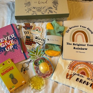Puede incluir: Un surtido de regalos que incluye un cuaderno rosa con el texto "Never Give Up", una agenda con un diseño de arcoíris, calcetines, pegatinas y un bálsamo labial con temática de piña. También hay una caja con el texto "Be kind".