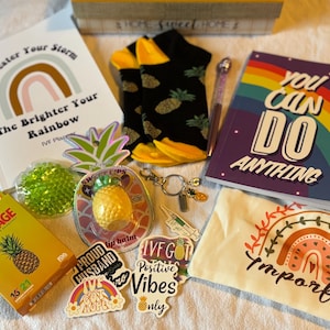 Puede incluir: Una colección de artículos con un tema positivo, que incluye un cuaderno con un diseño de arcoíris y las palabras "You Can Do Anything". Otros artículos incluyen pegatinas, un bálsamo labial, una venda y calcetines con estampados de piña.