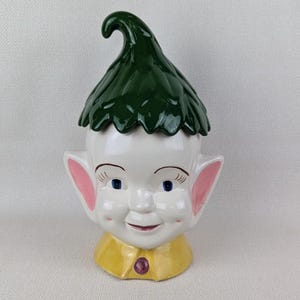 May include: A ceramic elf figurine with a green hat and a yellow shirt. The elf has a white face with blue eyes and a pink nose.
