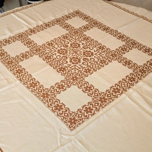 May include: A white tablecloth with a brown embroidered pattern of repeating floral designs. The tablecloth has a square design in the center and a border around the edges.