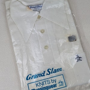 May include: A white polo shirt in a plastic package. The shirt has a large collar, three buttons, and a small pocket with a penguin logo. The packaging has the words "Grand Slam" and "KNITS by Munsingwear".