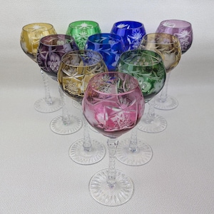 May include: A collection of ten colorful crystal wine glasses. Each glass features a different color, including pink, purple, green, blue, yellow, and gold. The glasses have intricate starburst patterns and clear stems and bases.