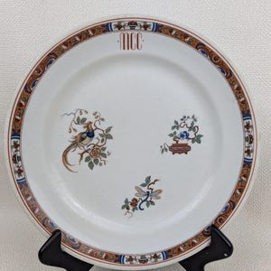 May include: White ceramic plate with a decorative border in brown, blue, and gold. The plate features a bird and floral design, with the letters "MCC" at the top. The plate is displayed on a black stand.