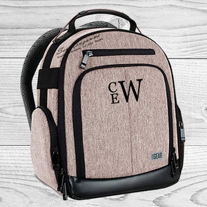 May include: A tan backpack with black accents, a top handle, and padded shoulder straps. The front features the initials "CW" and "E" in black. The backpack has multiple zippered compartments and the word "GEAR" is printed on the bottom right.