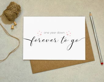 Peut inclure: Carte de vœux blanche avec l'inscription "one year down forever to go" en écriture noire. La carte est sur une enveloppe marron, avec un crayon et de la ficelle avec des punaises en arrière-plan. Petits cœurs rouges au-dessus des mots.