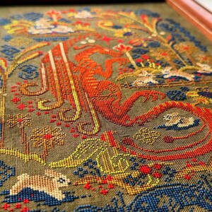 May include: Framed cross-stitch artwork featuring a detailed design with a large orange letter, possibly an initial. The piece includes floral and animal motifs in shades of blue, yellow, red, and green, set against a muted green background.