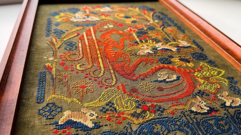 May include: Framed cross-stitch artwork featuring a detailed design with a large orange letter, possibly an initial. The piece includes floral and animal motifs in shades of blue, yellow, red, and green, set against a muted green background.