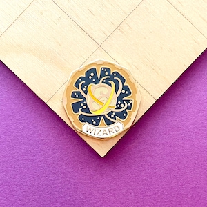以下が含まれることがあります： 惑星が渦巻く模様で囲まれた黒と黄色のデザインの金色のピン。ピンには「WIZARD」という文字が下部に書かれている。