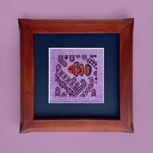 Può includere: Opera d'arte a punto croce incorniciata con un disegno di pesce. L'opera è ambientata su uno sfondo lavanda e incorniciata in una cornice di legno scuro. Il pesce è arancione, verde e rosso, con accenti viola e neri.