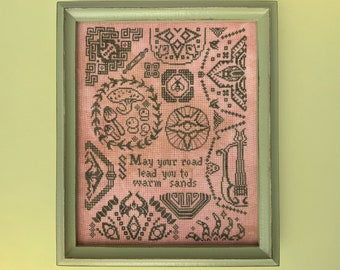 May include: A framed cross-stitch sampler with a pink background and black embroidery. The sampler features a variety of geometric designs and the text "May your road lead you to warm sands".