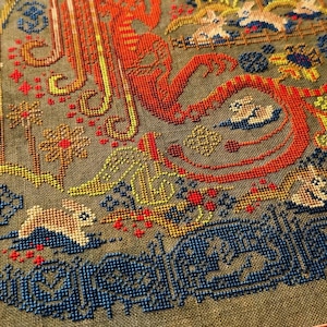 May include: Framed cross-stitch artwork featuring a detailed design with rabbits, flowers, and ornate patterns. The color palette includes shades of red, orange, yellow, blue, and green on a muted green background. The artwork is enclosed in a wooden frame.