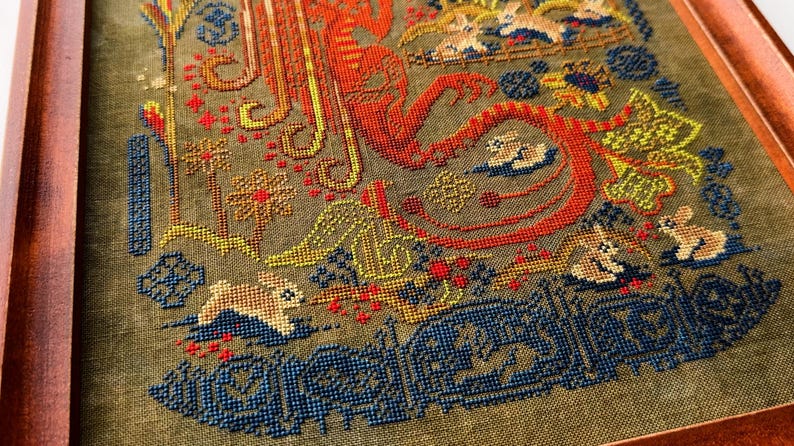 May include: Framed cross-stitch artwork featuring a detailed design with rabbits, flowers, and ornate patterns. The color palette includes shades of red, orange, yellow, blue, and green on a muted green background. The artwork is enclosed in a wooden frame.