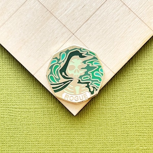 May include: A gold-colored pin with a skull design and the word "ROGUE" at the bottom. The skull is light green with dark green and teal accents. The pin is set against a light wood surface and a green background.