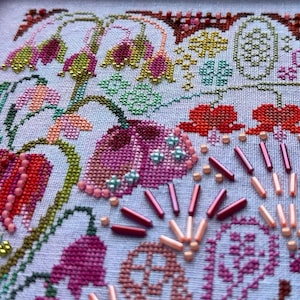 May include: A cross-stitch embroidery piece on light fabric featuring floral motifs in shades of pink, red, and green. The design is embellished with various small glass beads in pink, red, and light blue, arranged in patterns and scattered accents.