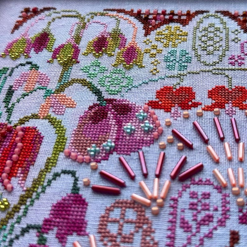 May include: A cross-stitch embroidery piece on light fabric featuring floral motifs in shades of pink, red, and green. The design is embellished with various small glass beads in pink, red, and light blue, arranged in patterns and scattered accents.