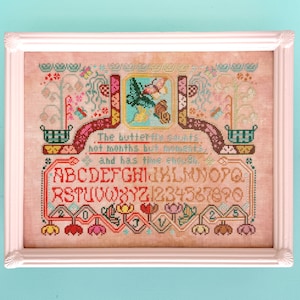 May include: Framed cross-stitch artwork featuring a detailed design with a butterfly motif. The piece includes an alphabet, numbers, and the quote "The butterfly counts not months but moments, and has time enough." The frame is white.