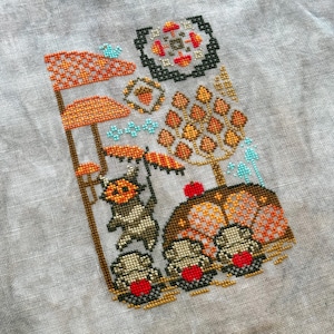 May include: Cross-stitch embroidery depicting a whimsical autumn scene with a small animal holding an umbrella, mushrooms, and a tree with orange leaves.