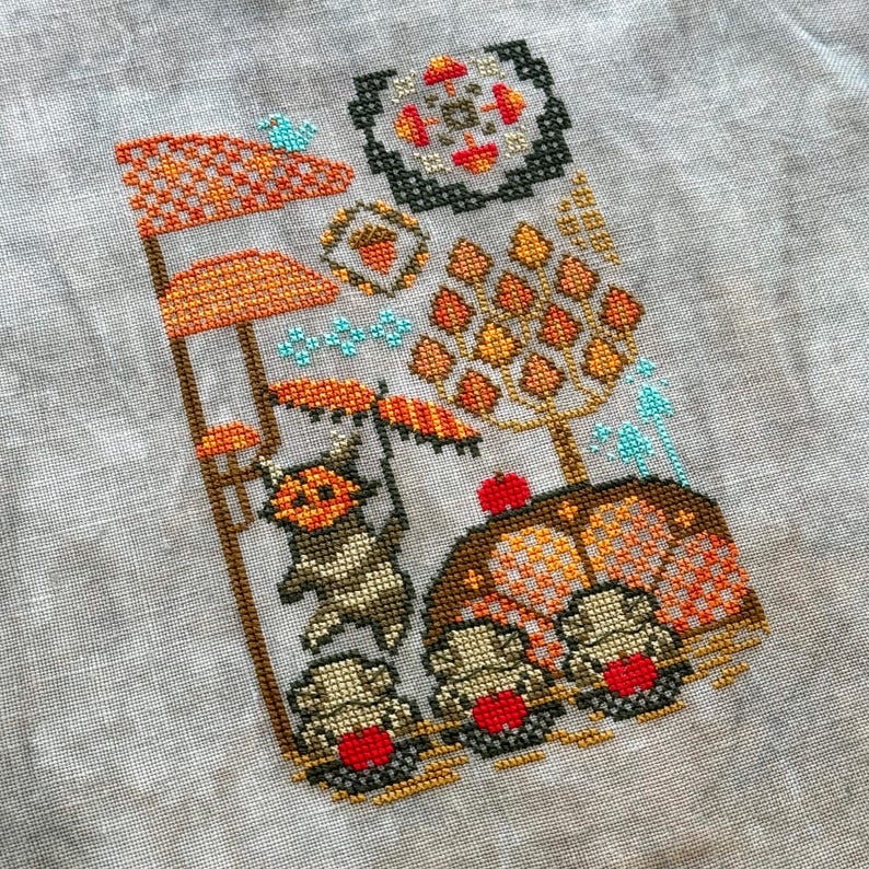 May include: Cross-stitch embroidery depicting a whimsical autumn scene with a small animal holding an umbrella, mushrooms, and a tree with orange leaves.