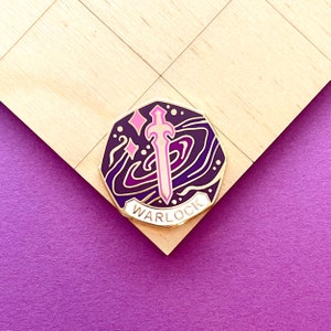 Puede incluir: Pin de esmalte dorado con un diseño morado y rosa. El pin presenta una espada con una hoja rosa y un fondo de galaxia en espiral. El texto "WARLOCK" está escrito en blanco sobre una pancarta blanca.