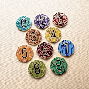 May include: A set of ten numbered enamel pins in various colours. Each pin has a unique design and colour scheme, with numbers from zero to nine. The pins are hexagonal and could be used for collecting or as decorative accessories.