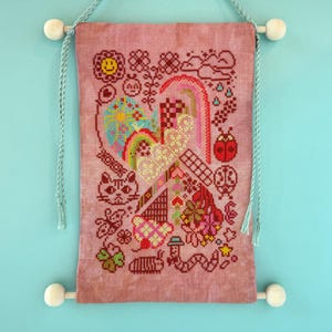 May include: A cross-stitch wall hanging with a pink background. The design features a heart, rainbow, cat, ladybug, and other whimsical elements. The hanging is suspended by wooden dowels and light blue cords.