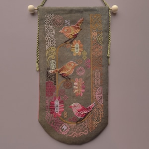 May include: A decorative wall hanging featuring cross-stitch embroidery. The design includes three birds perched on floral branches, with a color palette of brown, pink, and green. The banner is suspended by a wooden dowel and a green cord.
