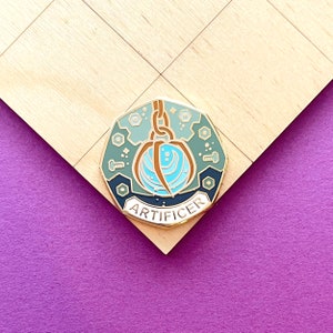 May include: A teal, gold, and blue enamel pin with the word "ARTIFICER" at the bottom. The pin features a stylized design of a chain, a sphere, and other geometric shapes. The pin is set against a wooden surface.